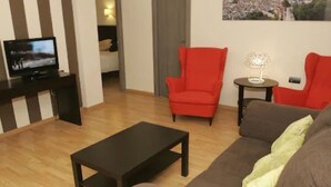 Una sala de estar con un sofá marrón, un sillón rojo, una mesa baja negra, un televisor sobre un mueble bajo y una mesita con una lámpara.