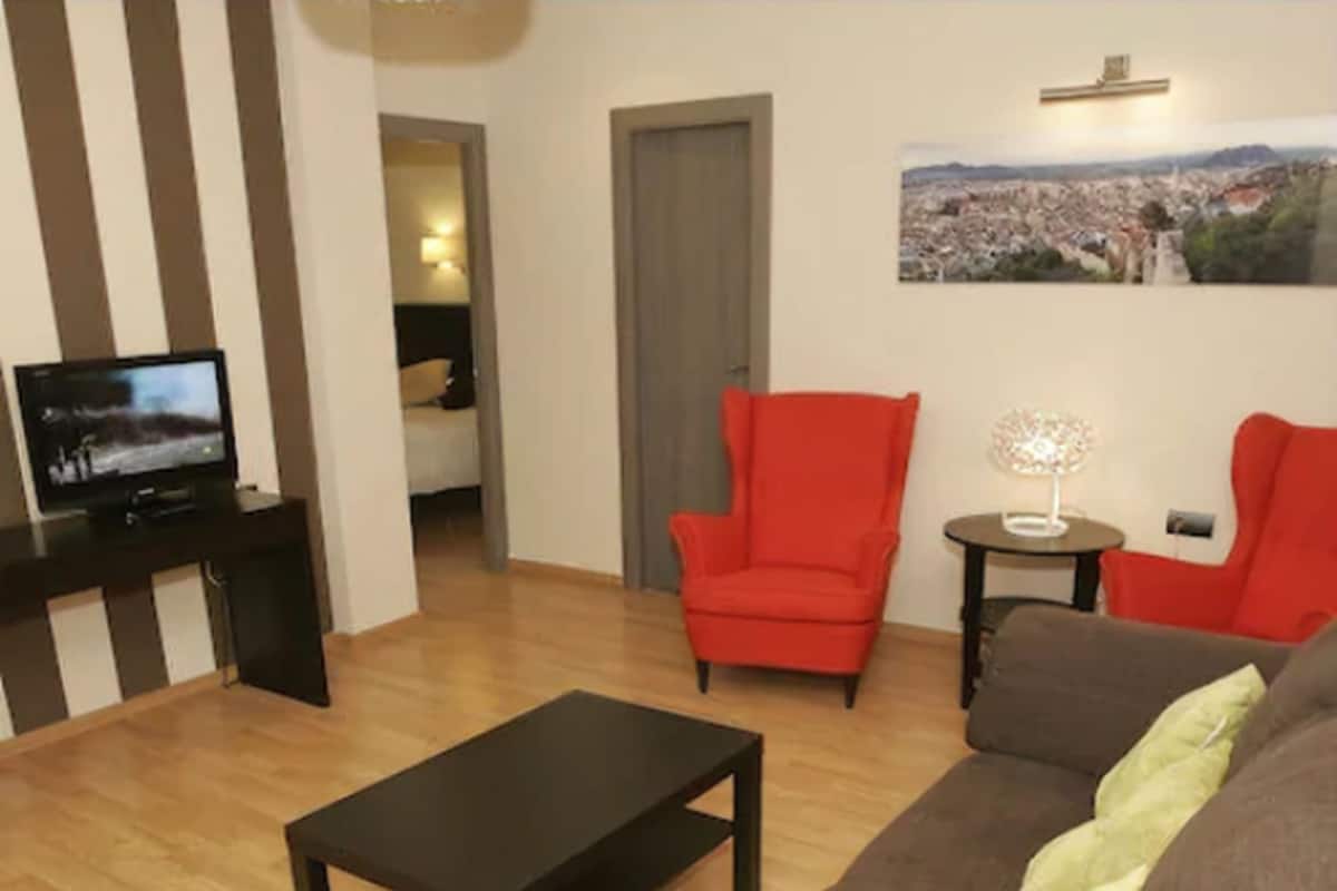 Ein Wohnzimmer mit einem braunen Sofa, einem roten Sessel, einem schwarzen Couchtisch, einem Fernseher auf einem Schrank und einem kleinen Tisch mit einer Lampe.