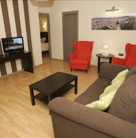 Una sala de estar con un sofá marrón, un sillón rojo, una mesa baja negra, un televisor sobre un mueble bajo y una mesita con una lámpara.