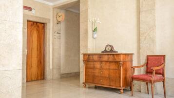 Een houten dressoir met laden, een rode fauteuil, een klok en een vaas met bloemen aan de muur.