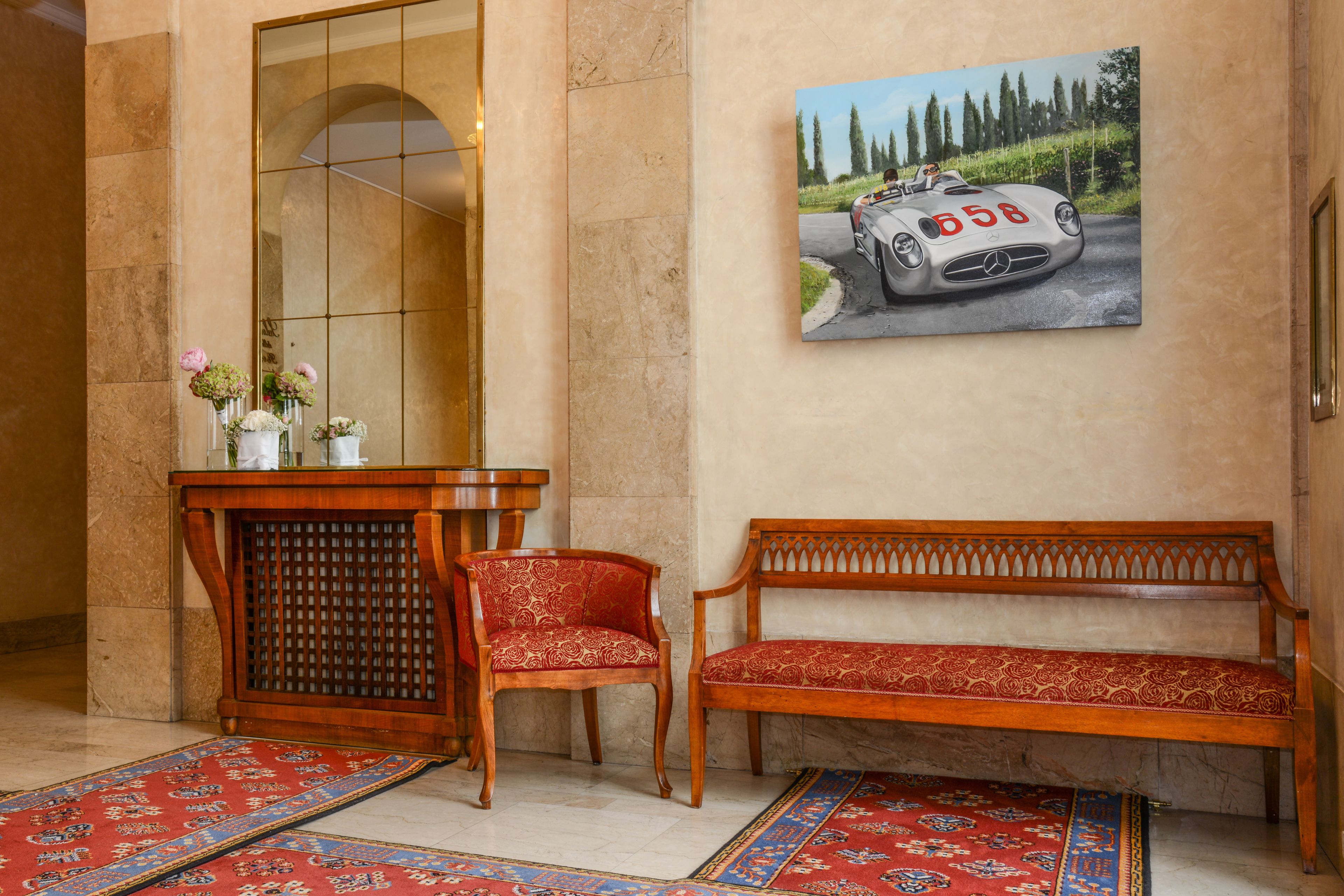 Un hall d’entrée avec un banc en bois, un canapé rouge à motifs, un comptoir d’accueil en bois et un tableau représentant une voiture accroché au mur.