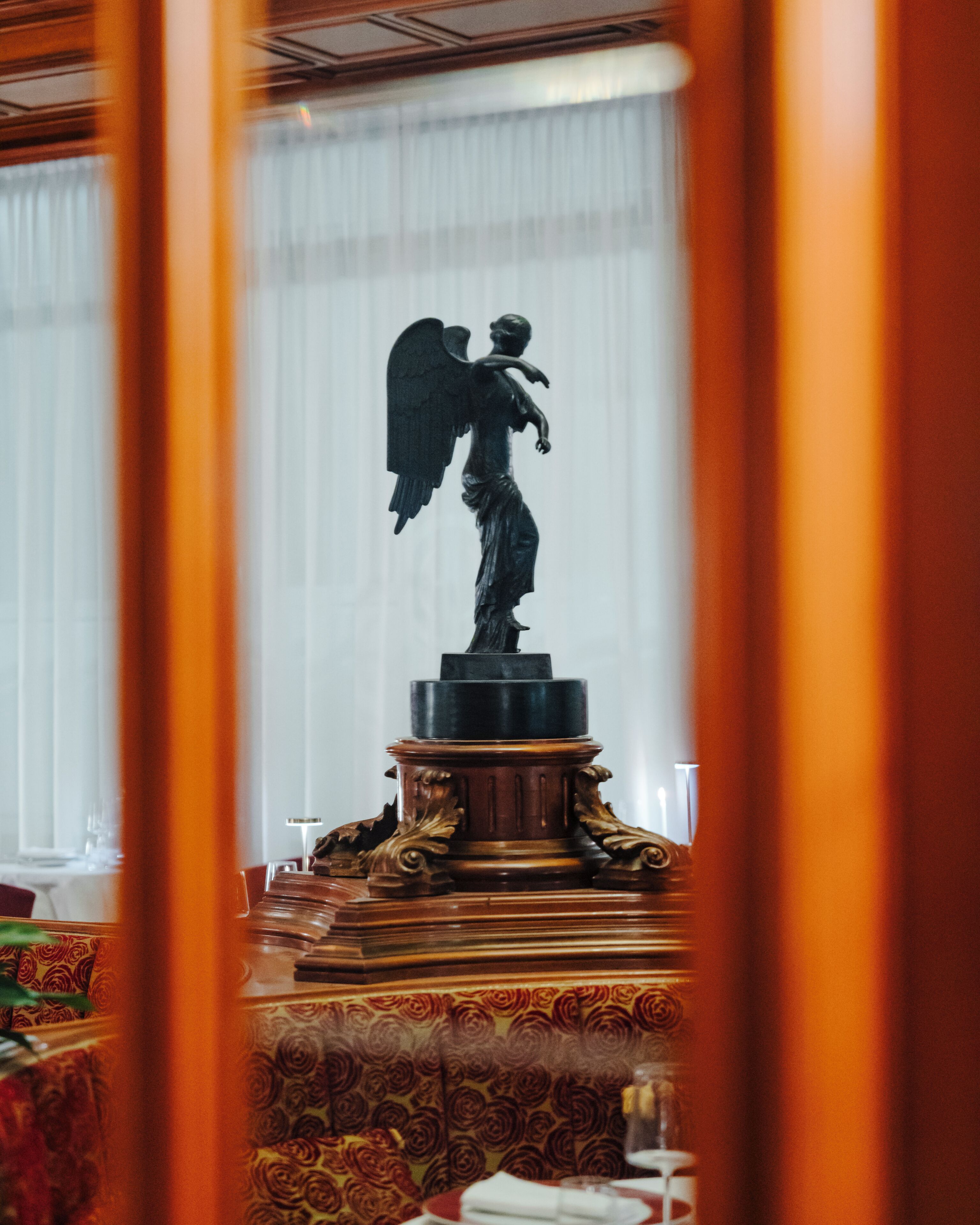 A statue of an angel on a pedestal in a room with patterned carpet and a table with a glass.