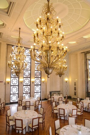 Une salle de banquet dotée de nombreuses tables rondes dressées pour le repas, de lustres somptueux et de grandes fenêtres ornées de panneaux de verre décoratifs.