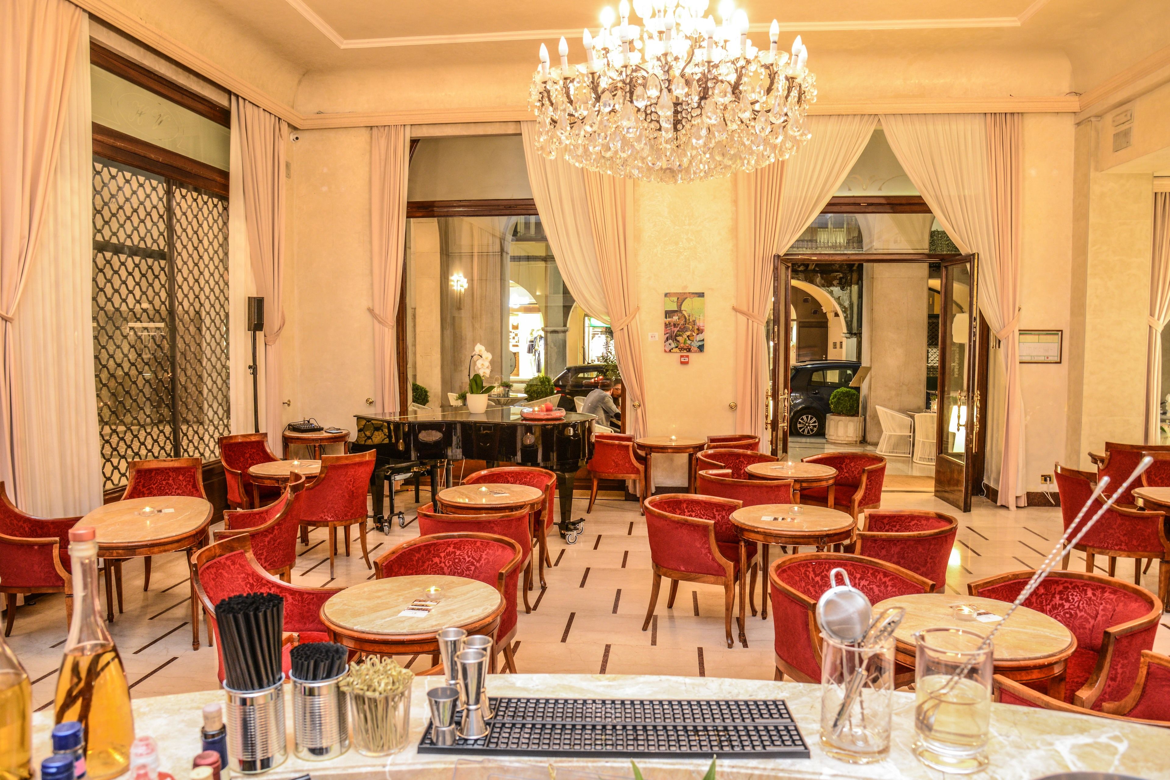 Eine Hotellobby mit runden Tischen, rot gepolsterten Sesseln, einer Bar und einer Kristallleuchtre.'