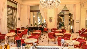 Eine Hotellobby mit runden Tischen, rot gepolsterten Sesseln, einer Bar und einer Kristallleuchtre.'