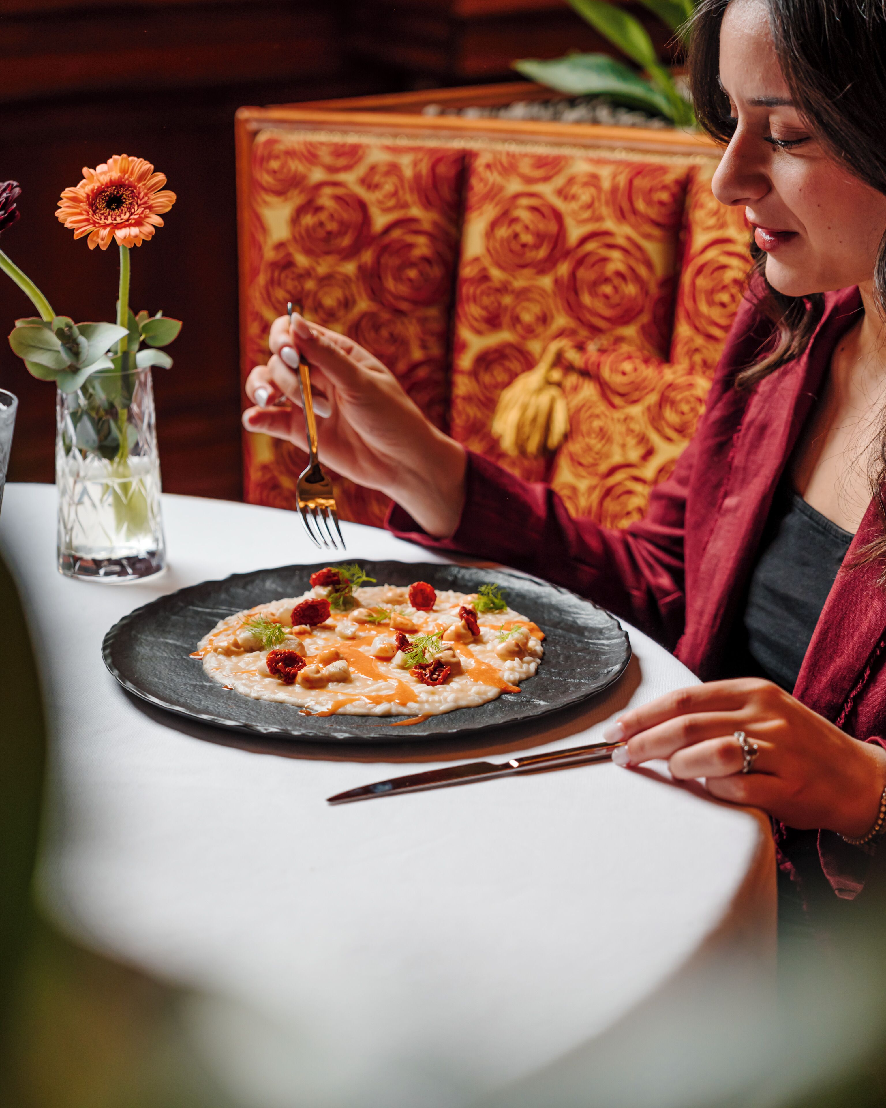 A dining table with a plate of food, a vase with flowers, and a person holding a fork.
