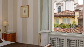 Camera d'albergo con un letto grande, pavimento in legno, un lampadario e una finestra con vista su un edificio storico.