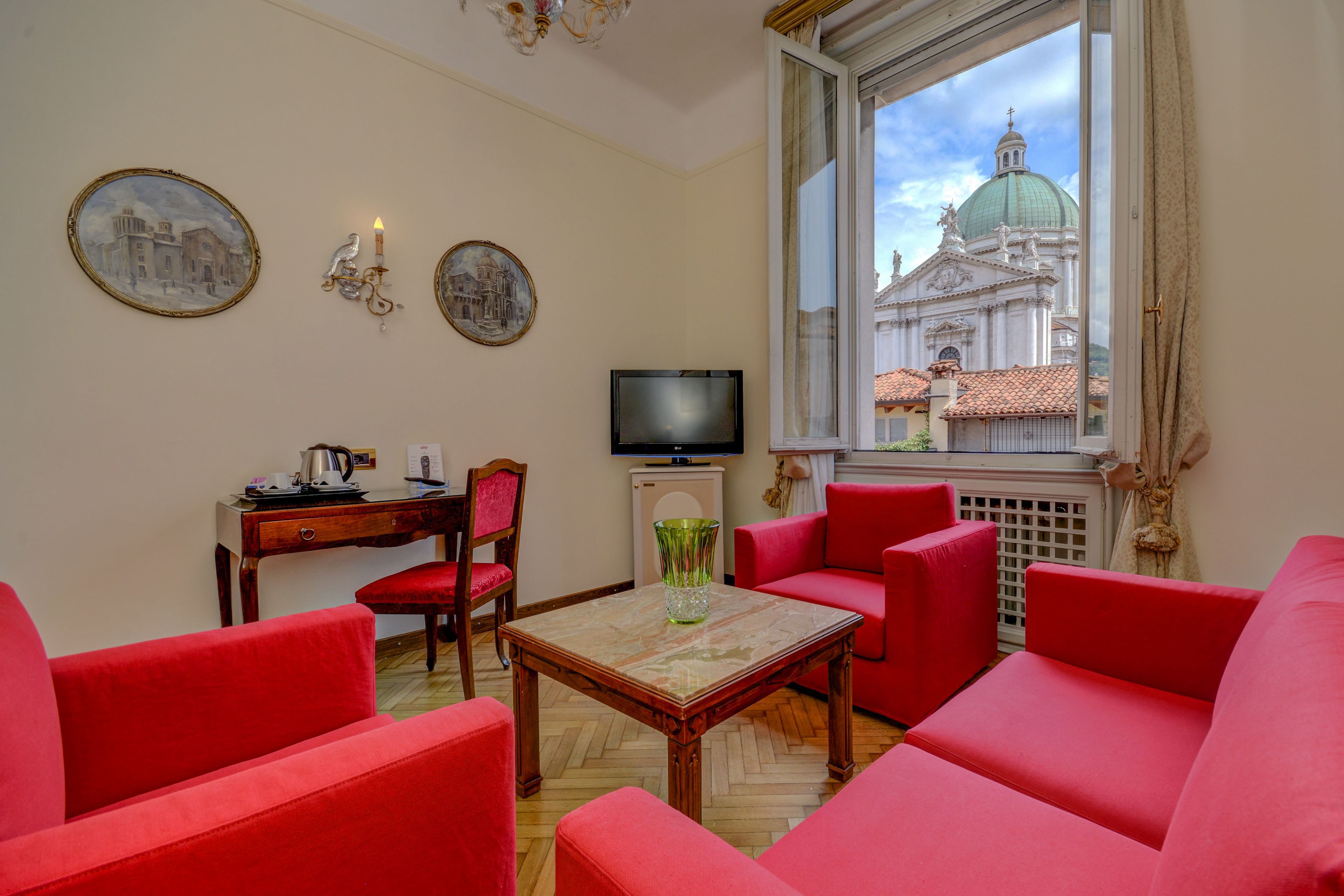 A room with red sofas, a wooden table, a TV, and a window with a view of a building with a dome.