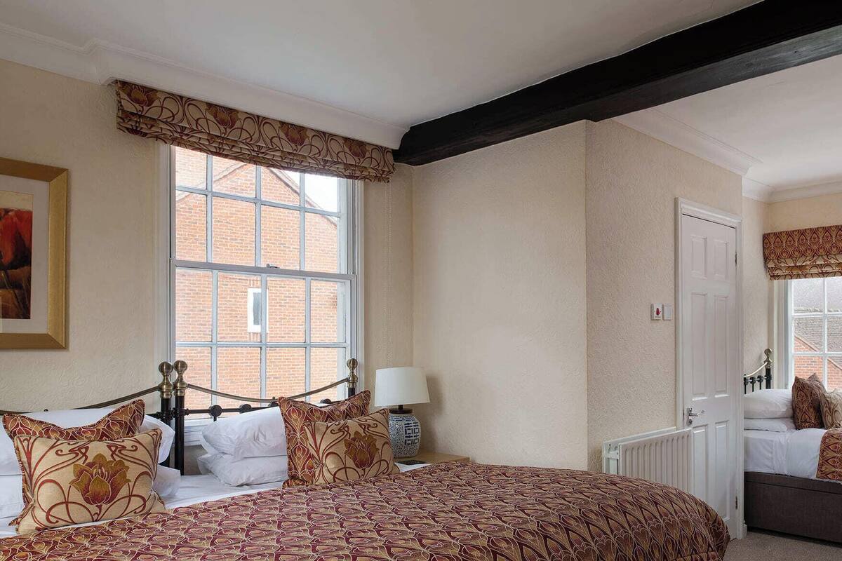 A bedroom with a large window, a bed with patterned bedding, a bedside table with a lamp, and a radiator.