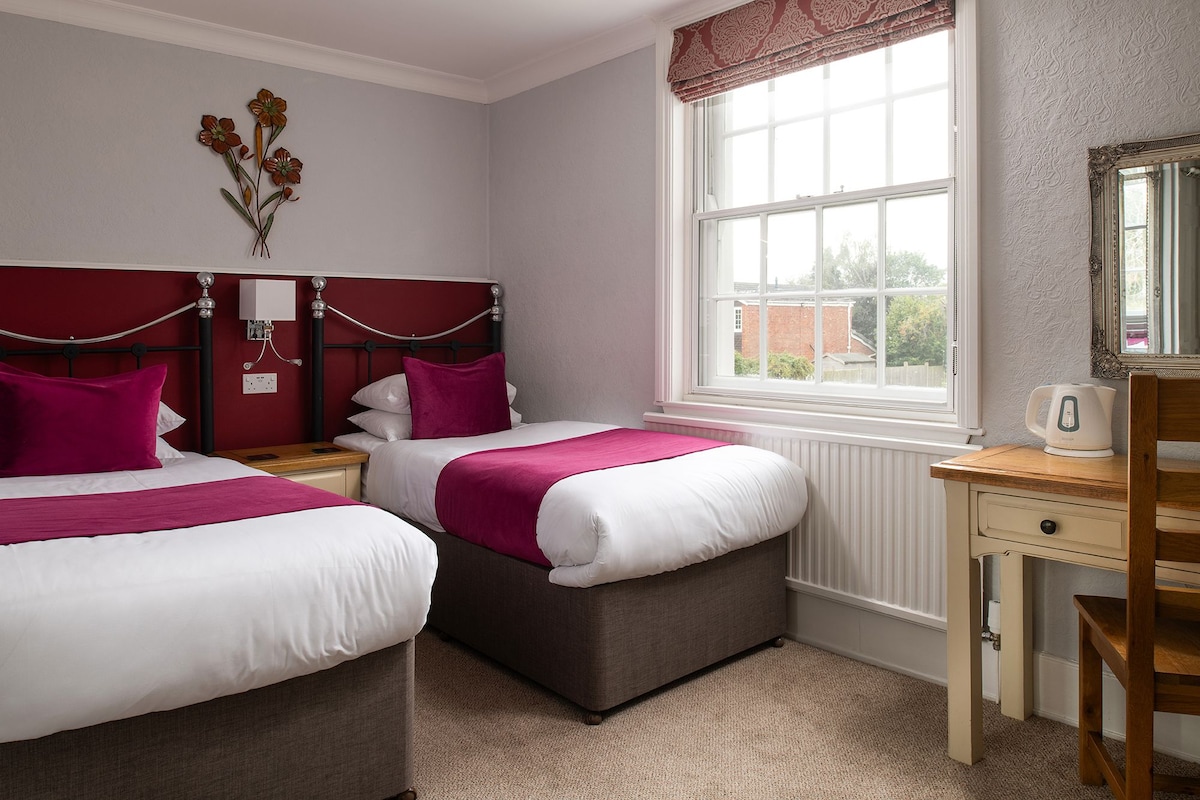 A hotel room with two beds, a wooden desk, a radiator, and a window with a view of the outside.