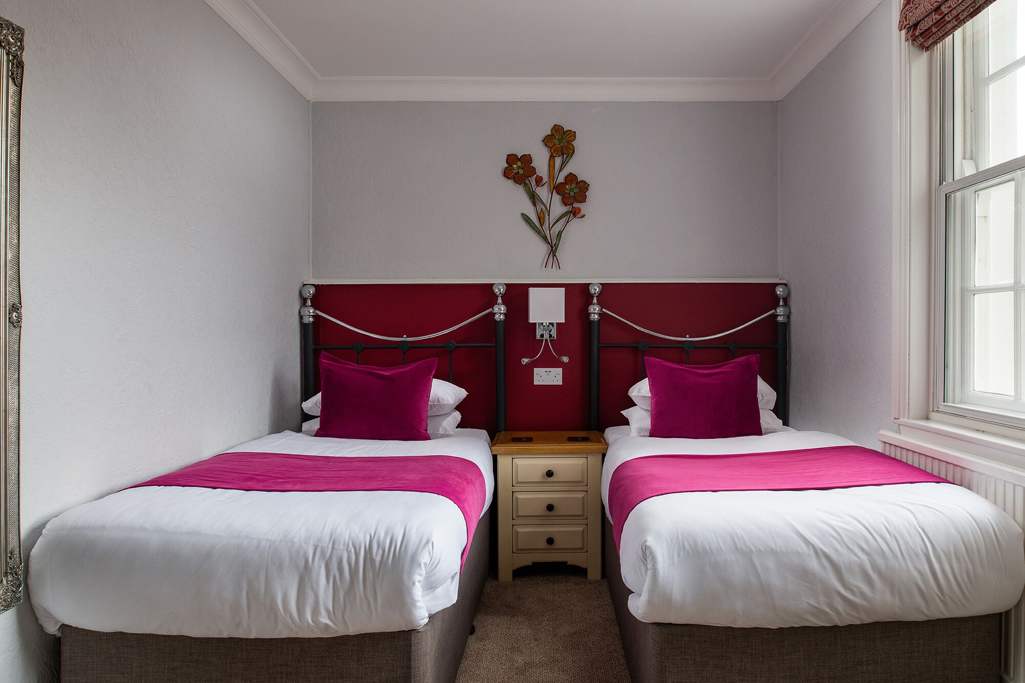 Habitación doble con dos camas, cada una con cabecero rojo y ropa de cama blanca. Entre las camas hay una mesita de noche de madera, y una lámpara fijada a la pared arriba.