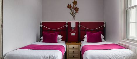 Habitación doble con dos camas, cada una con cabecero rojo y ropa de cama blanca. Entre las camas hay una mesita de noche de madera, y una lámpara fijada a la pared arriba.