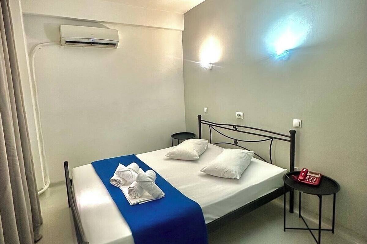A single bed with a blue bedspread, white pillows, and a rolled towel. There is a small bedside table with a red telephone and a wall-mounted light. An air conditioning unit is visible on the wall.