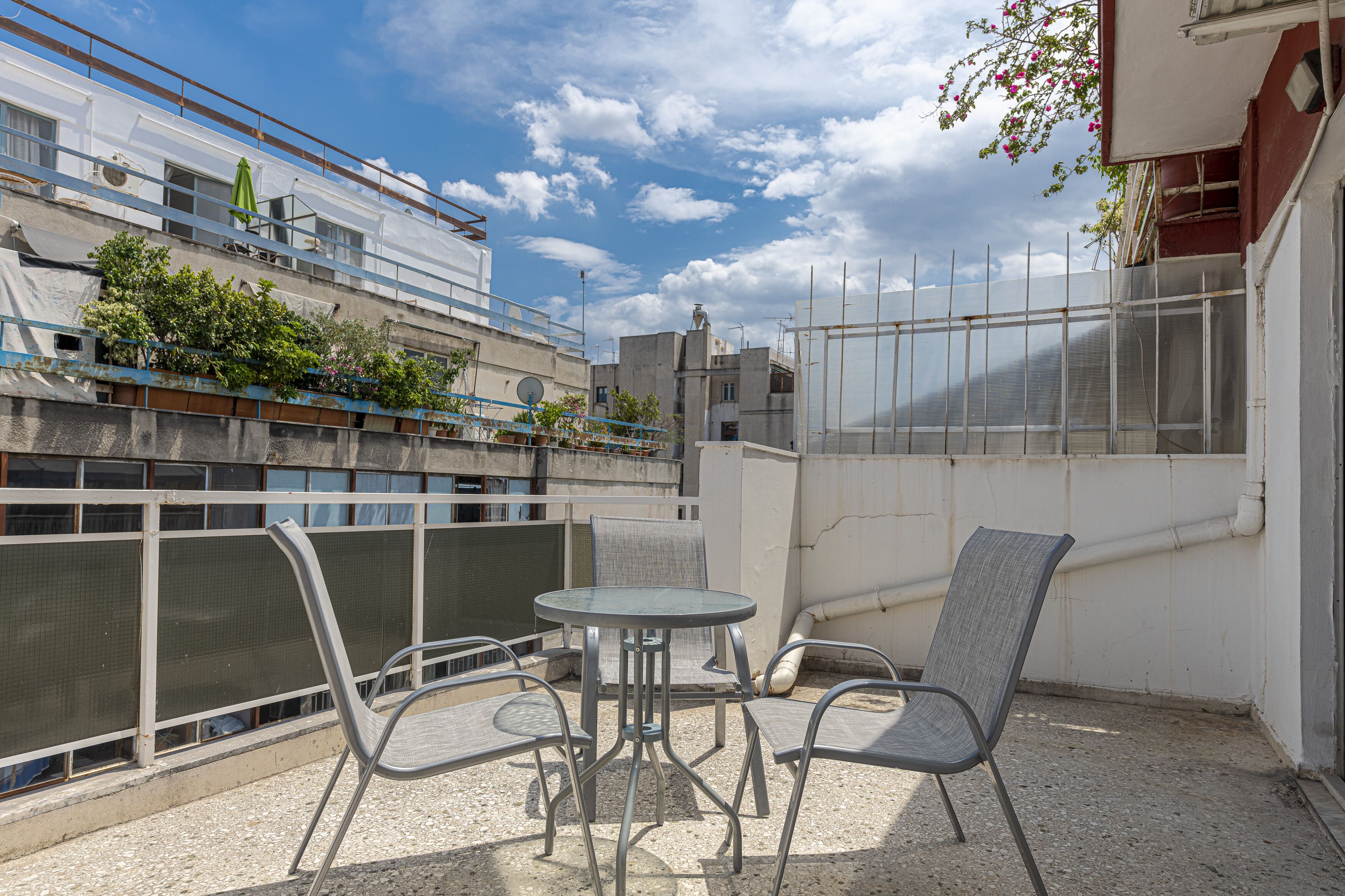 A balcony with a metal table and chairs, a view of neighboring buildings, and a clear sky.