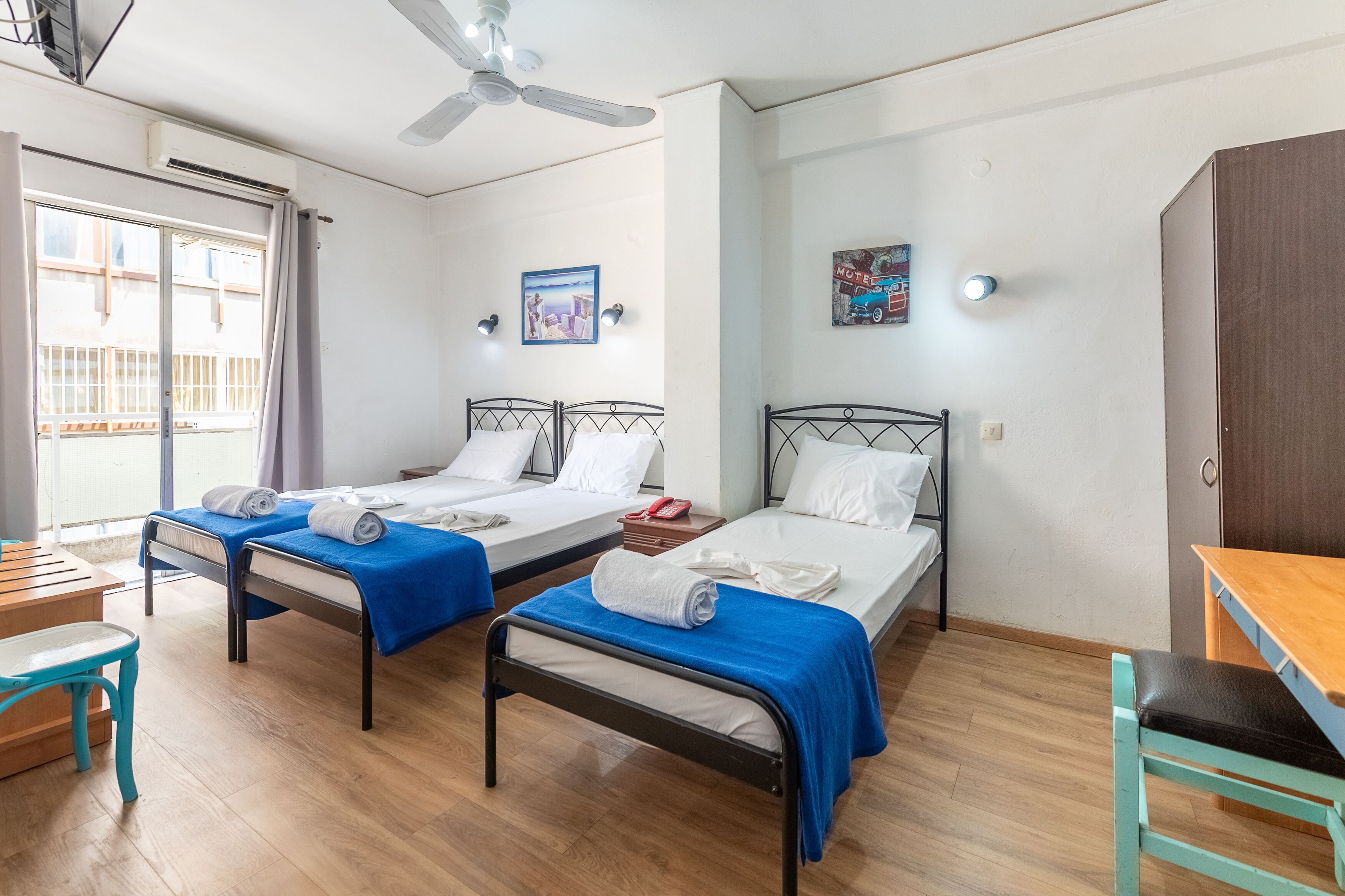 Habitación de hotel con dos camas individuales, un escritorio de madera, una silla, un ventilador de techo y una ventana con cortinas.