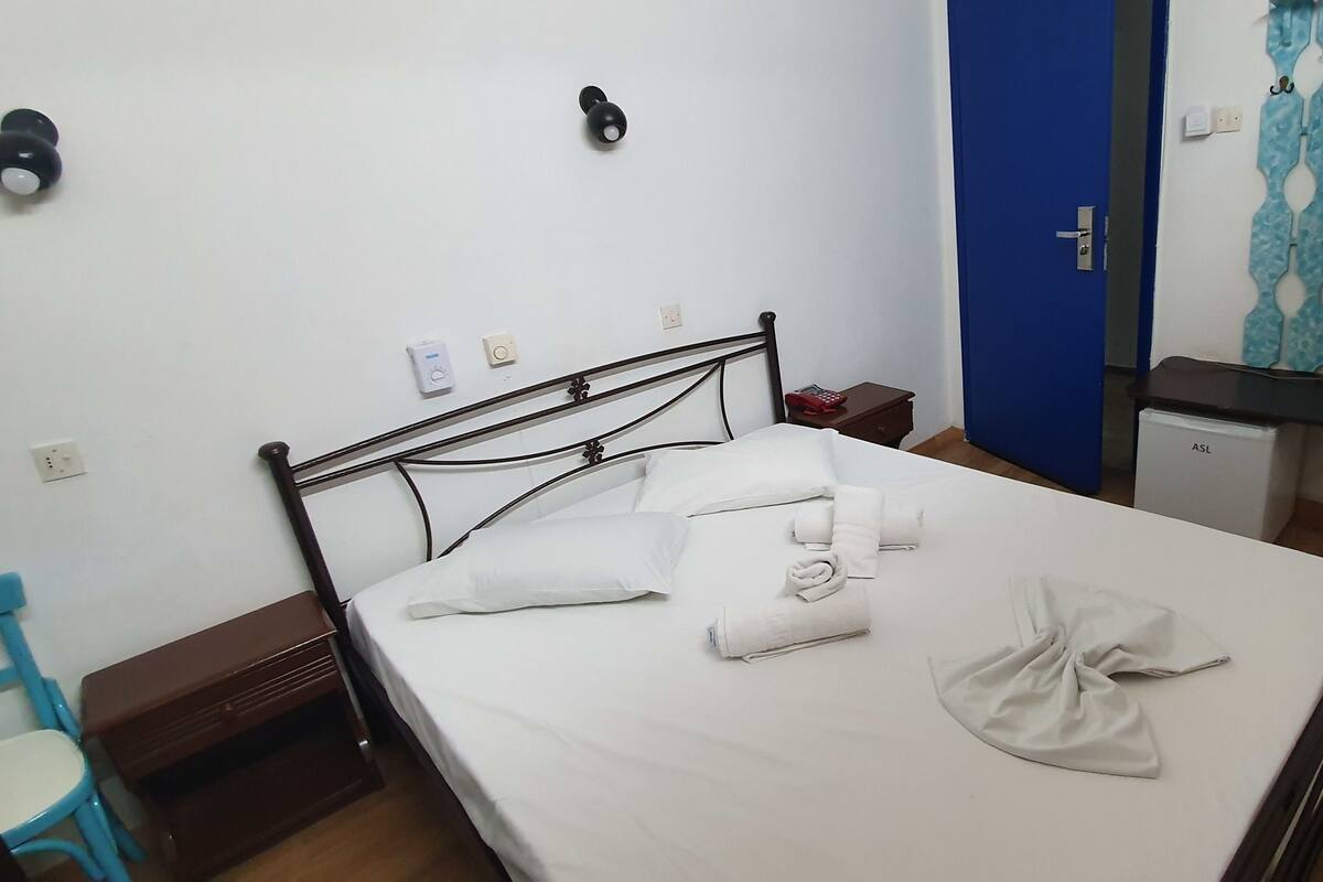 A neatly made bed with white linens and towels, a blue chair, a small bedside table, and a wall-mounted light fixture.