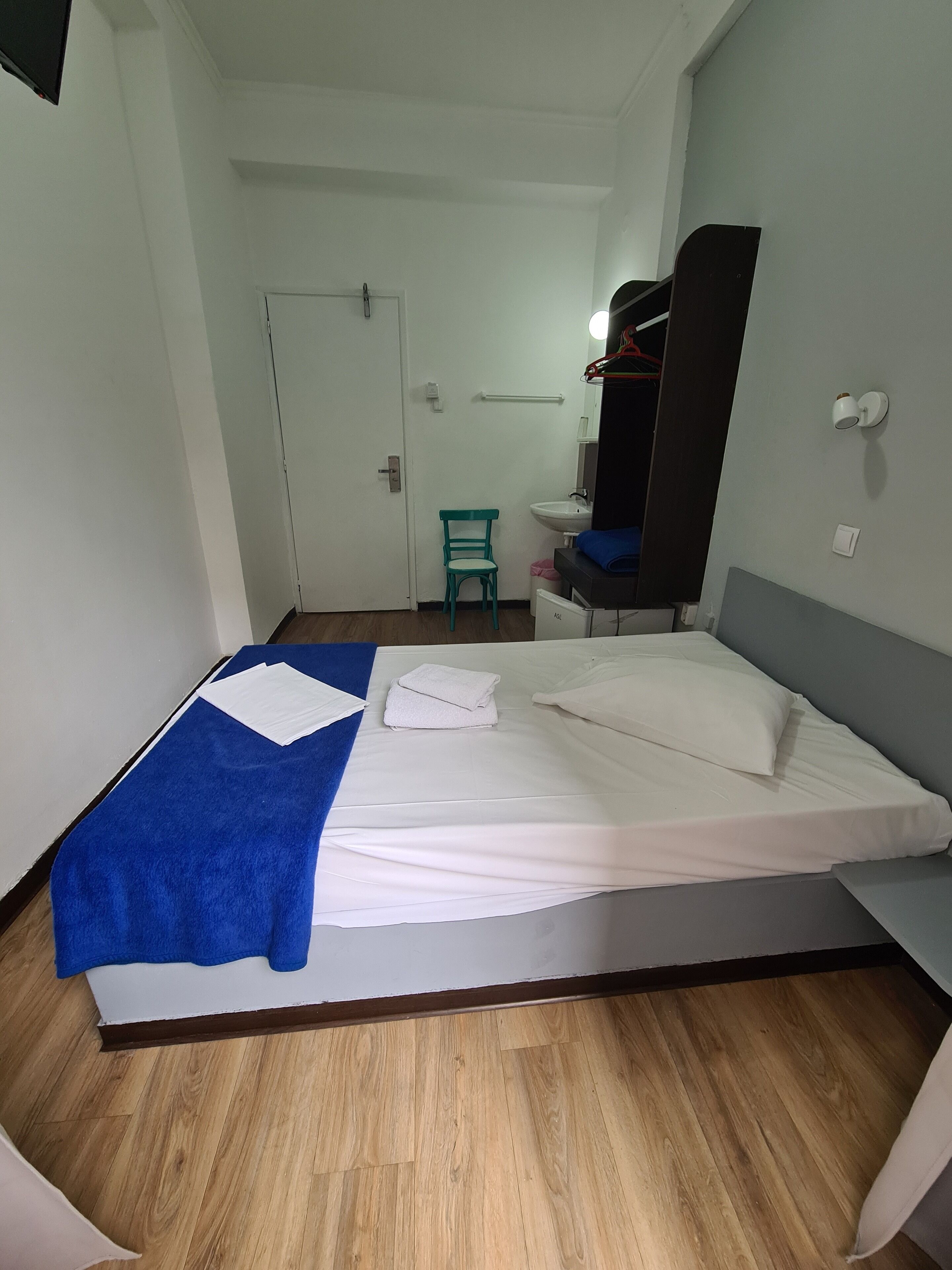 Une chambre d’hôtel avec un lit, une serviette bleue, un oreiller blanc, une petite chaise, un lavabo et un miroir.