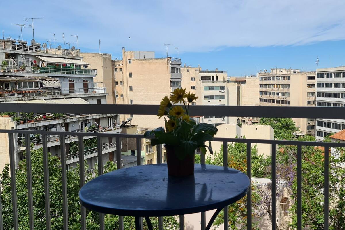 A balcony with a potted plant and a round table, overlooking a cityscape with multiple buildings.