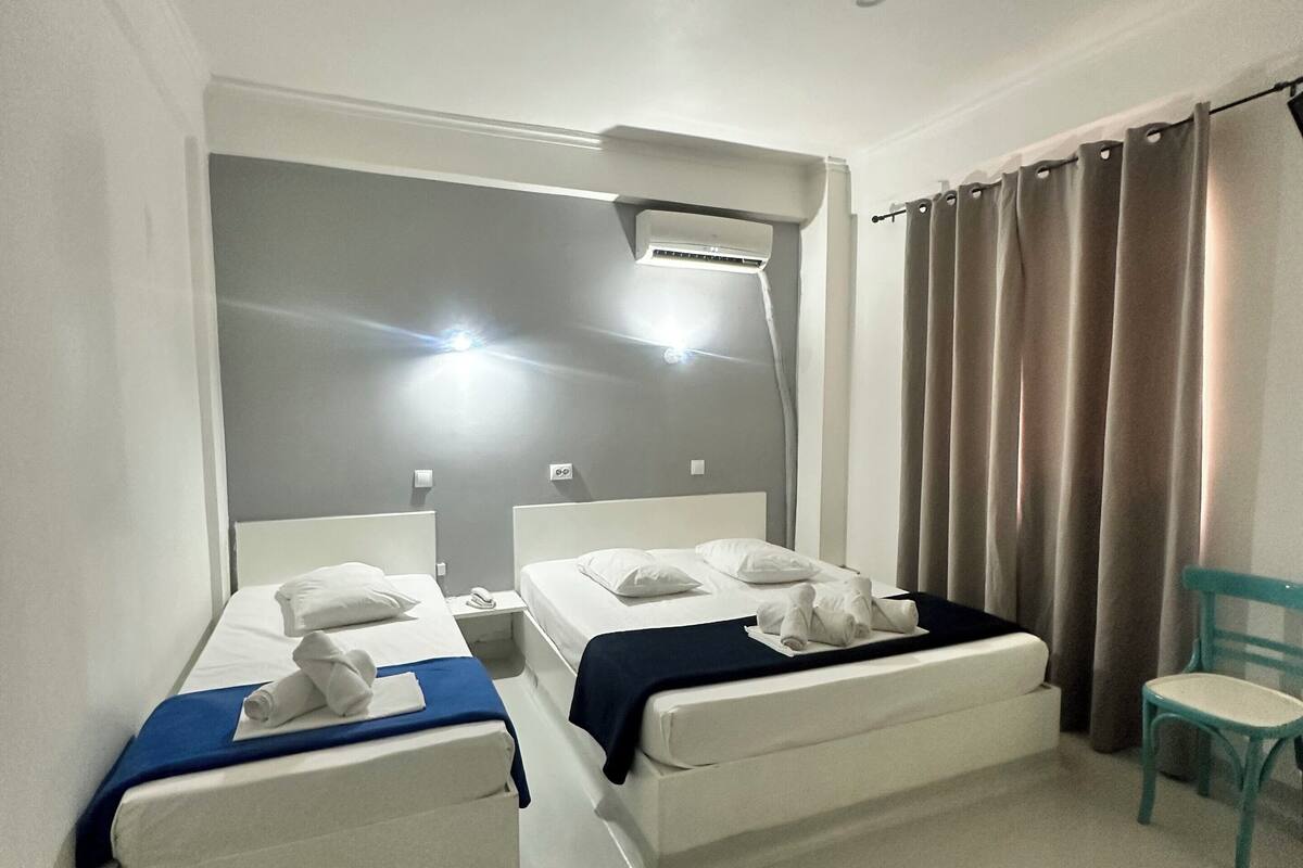 A hotel room with two beds, a ceiling light, an air conditioning unit, a chair, and a curtain.