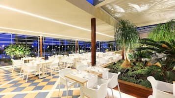 Un restaurante con mesas y sillas blancas, piso a cuadros y plantas de interior.