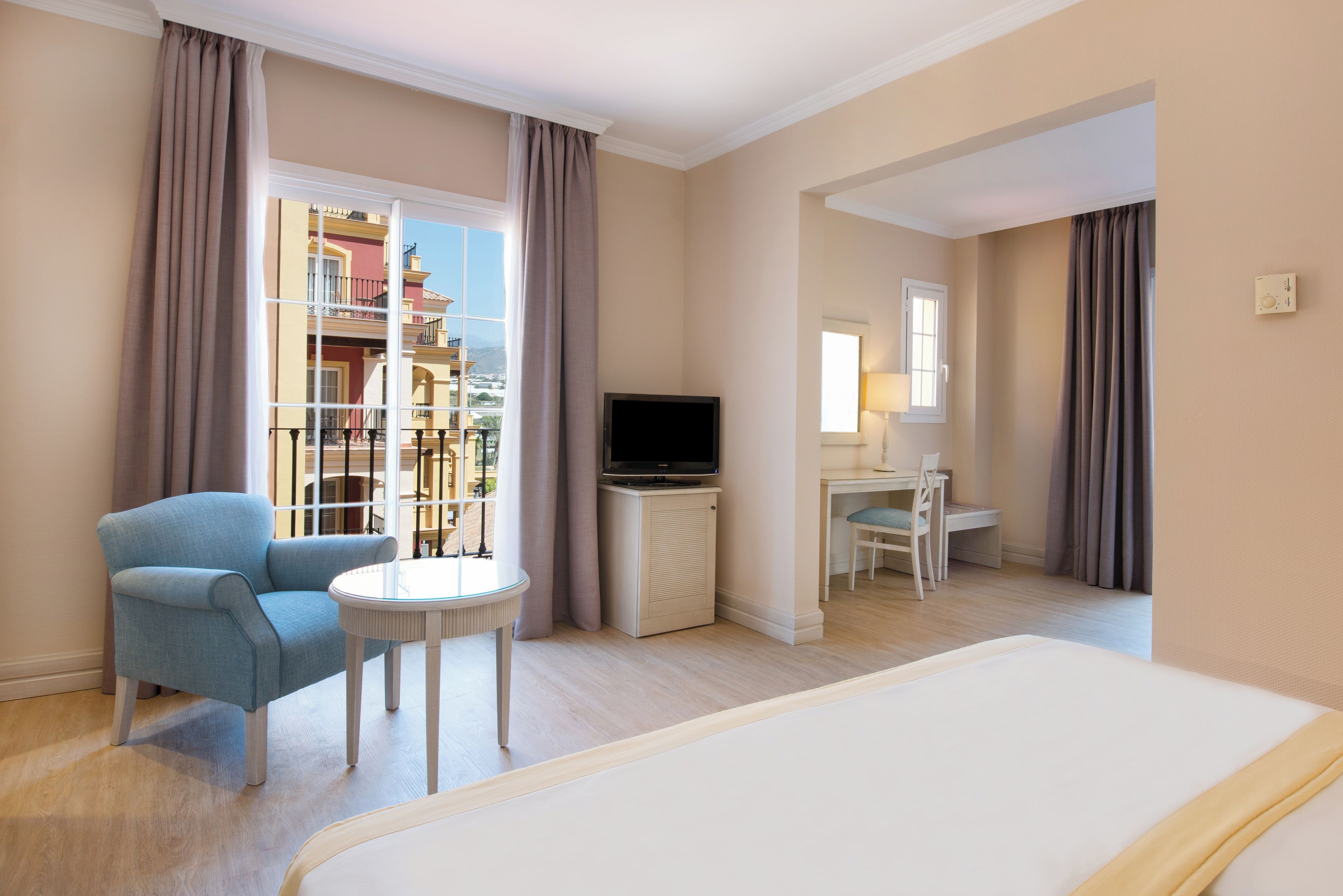 Una habitación de hotel con cama, escritorio, silla, televisor y vistas a un edificio a través del ventanal.