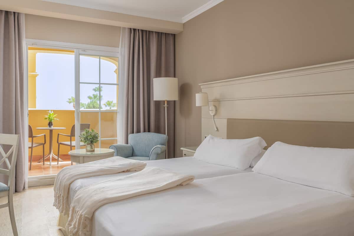 Quarto de hotel com uma cama grande, uma mesa redonda pequena com cadeiras, uma poltrona azul e vista para o exterior através de uma janela ampla.