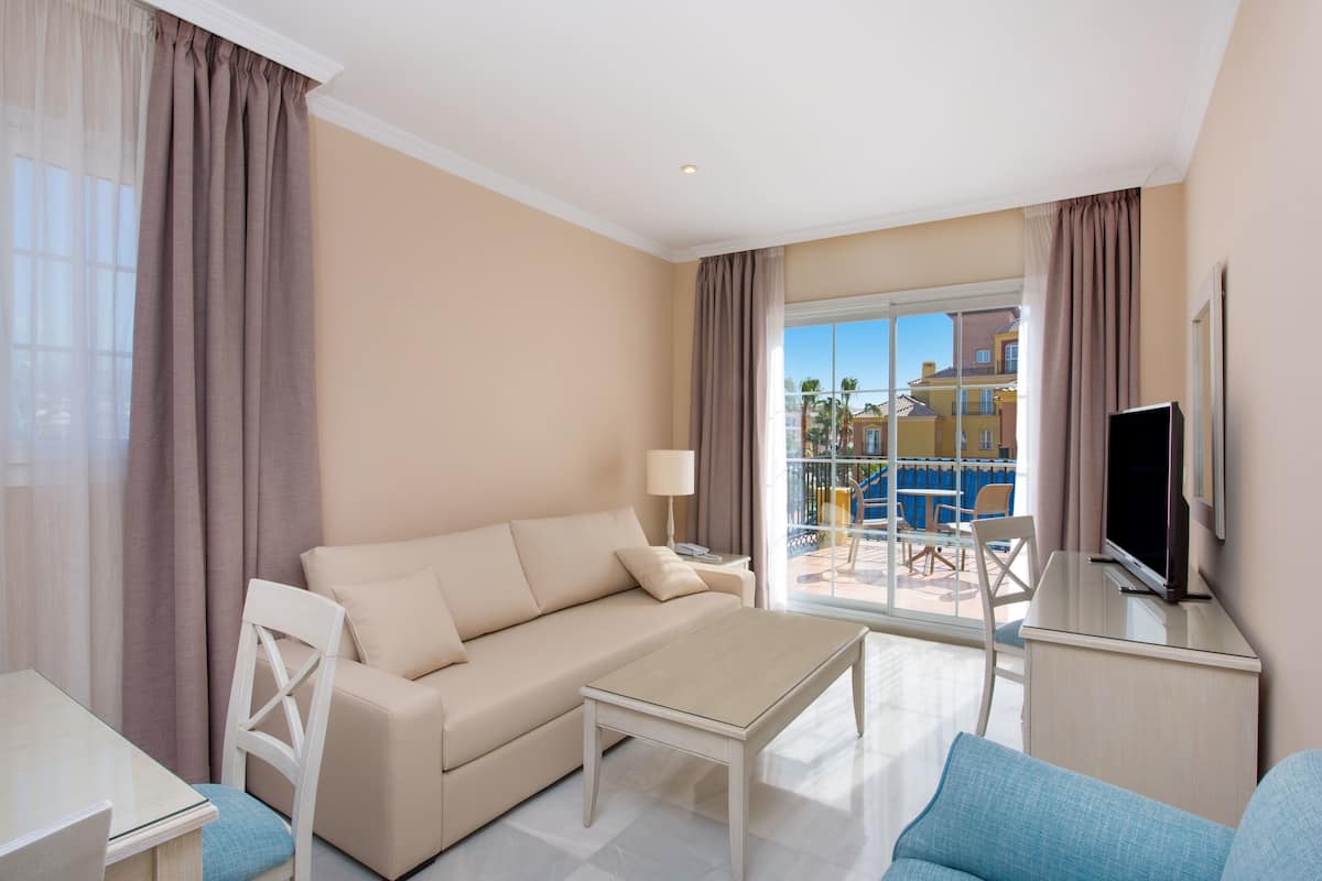 Quarto de hotel moderno com sofá, mesa de centro, TV e varanda com mobiliário exterior.