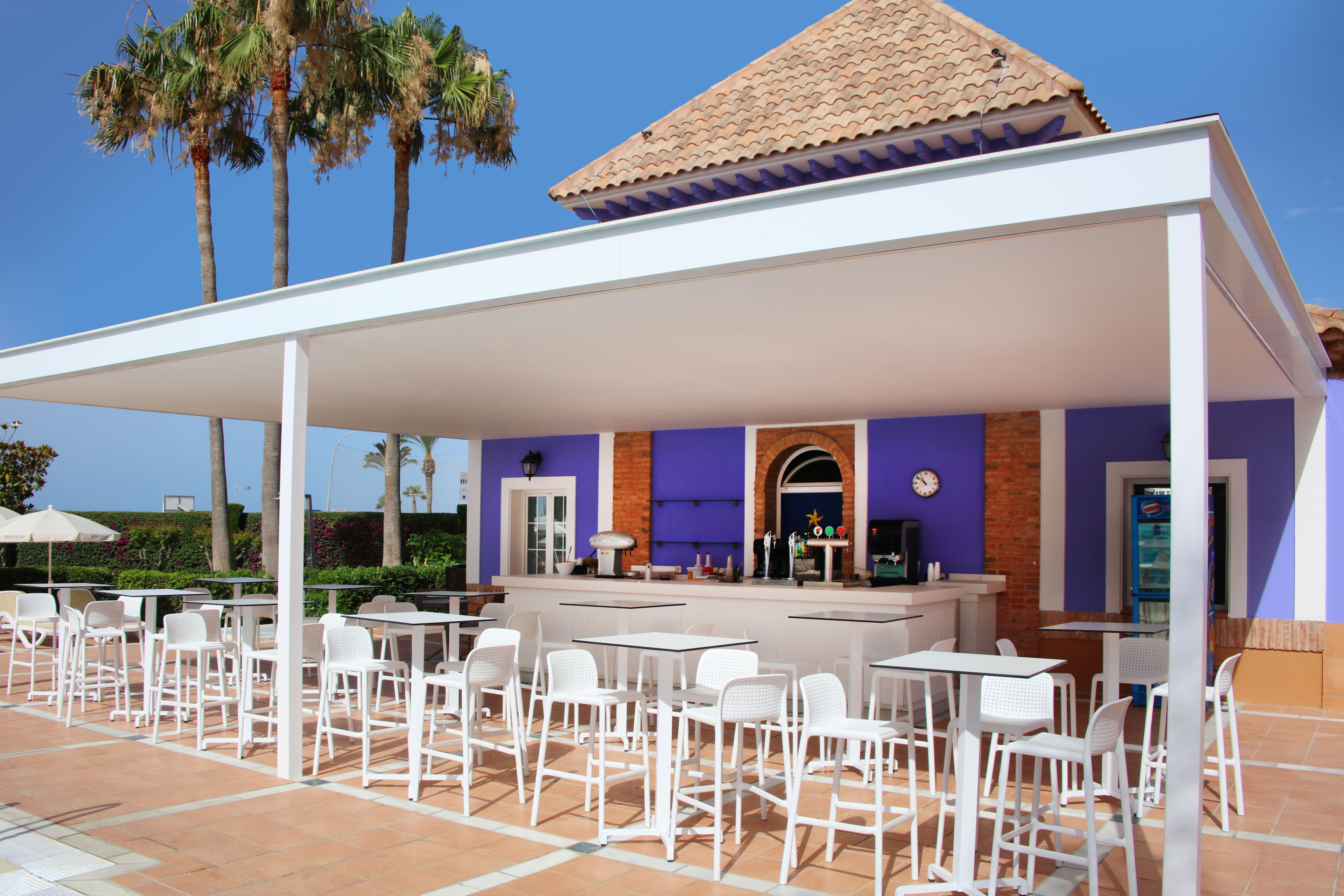 Un patio al aire libre con mesas y sillas blancas, una zona de estar cubierta y un bar con fachada morada y de ladrillo.