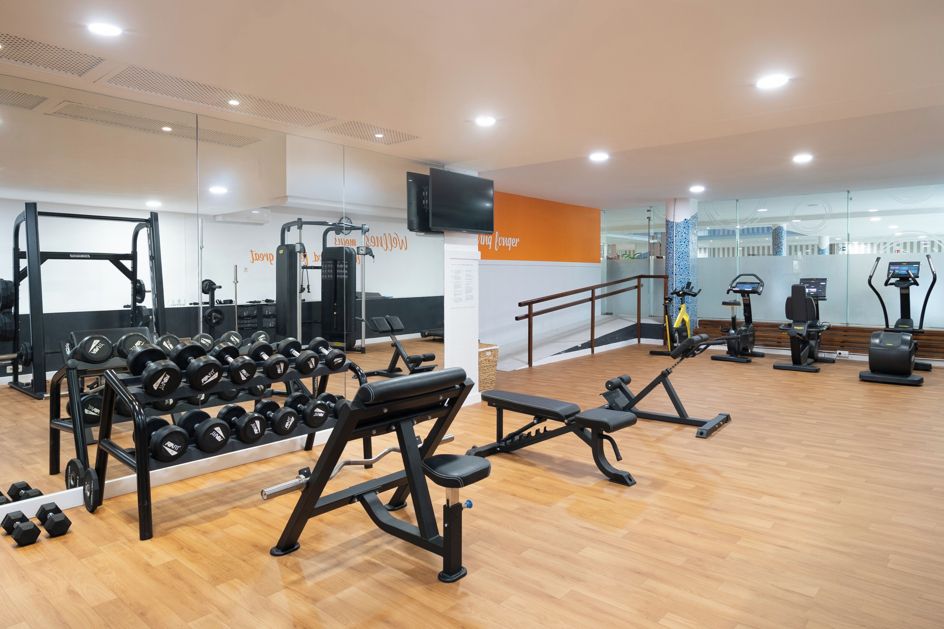 A gym with various exercise equipment, a television mounted on the wall, and a wooden floor.