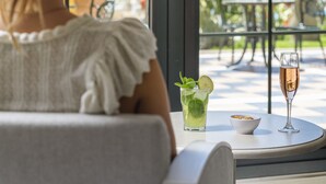 Eine Frau sitzt an einem Tisch mit einem Getränk und einer Schüssel und blickt auf eine Terrasse mit Gartenmöbeln und Palmen.