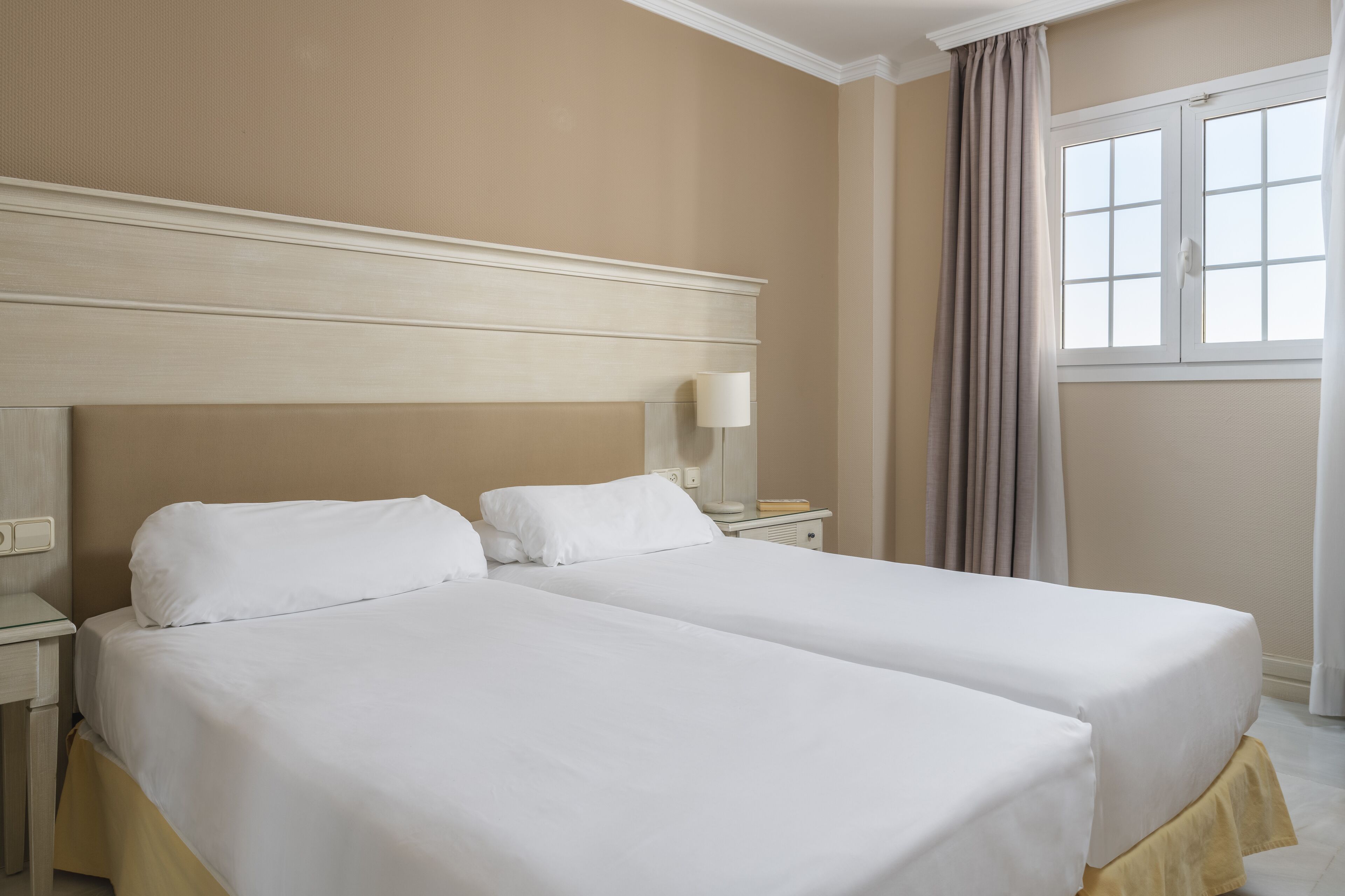 A hotel room with two single beds, a nightstand with a lamp, a window with curtains, and a beige wall.