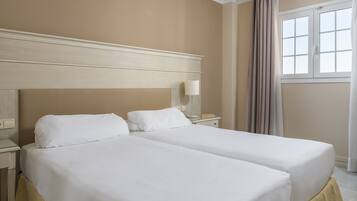 A hotel room with two single beds, a nightstand with a lamp, a window with curtains, and a beige wall.
