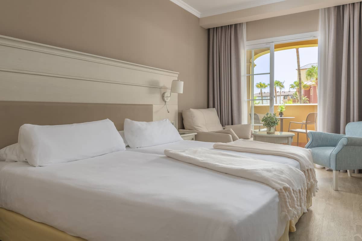 Quarto de hotel com uma cama grande, uma cadeira, uma pequena mesa com uma planta e vista para o exterior.