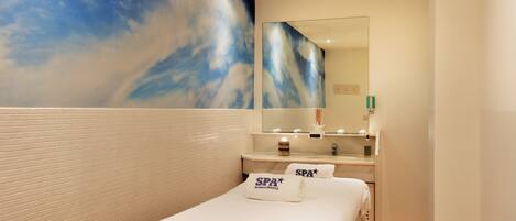 Habitació d'hotel amb un llit, un mirall gran, una tocador amb un lavabo i una paret amb un mural de núvols.