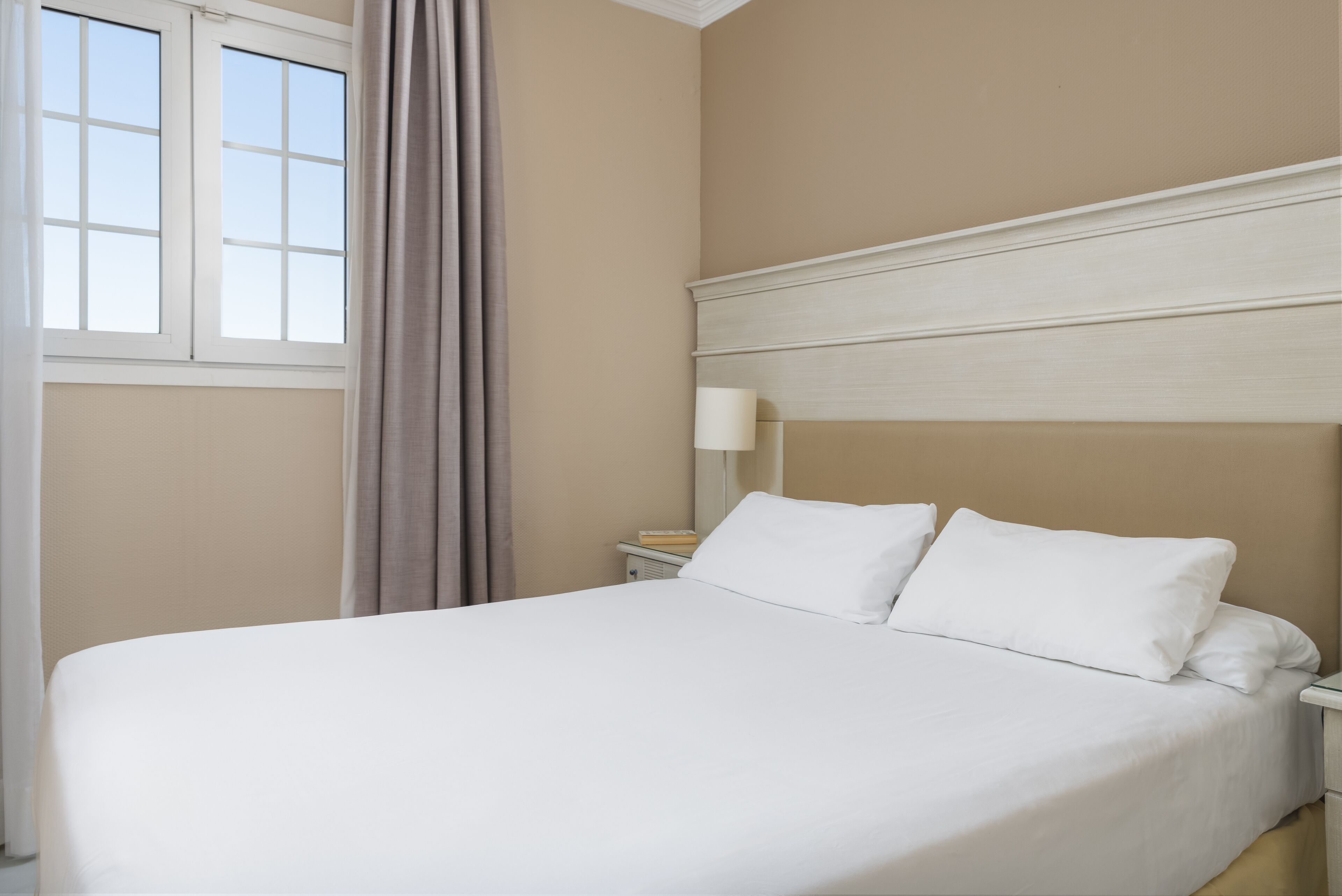 Een net opgemaakt bed met witte lakens in een kamer met beige wanden en een raam met gordijnen.