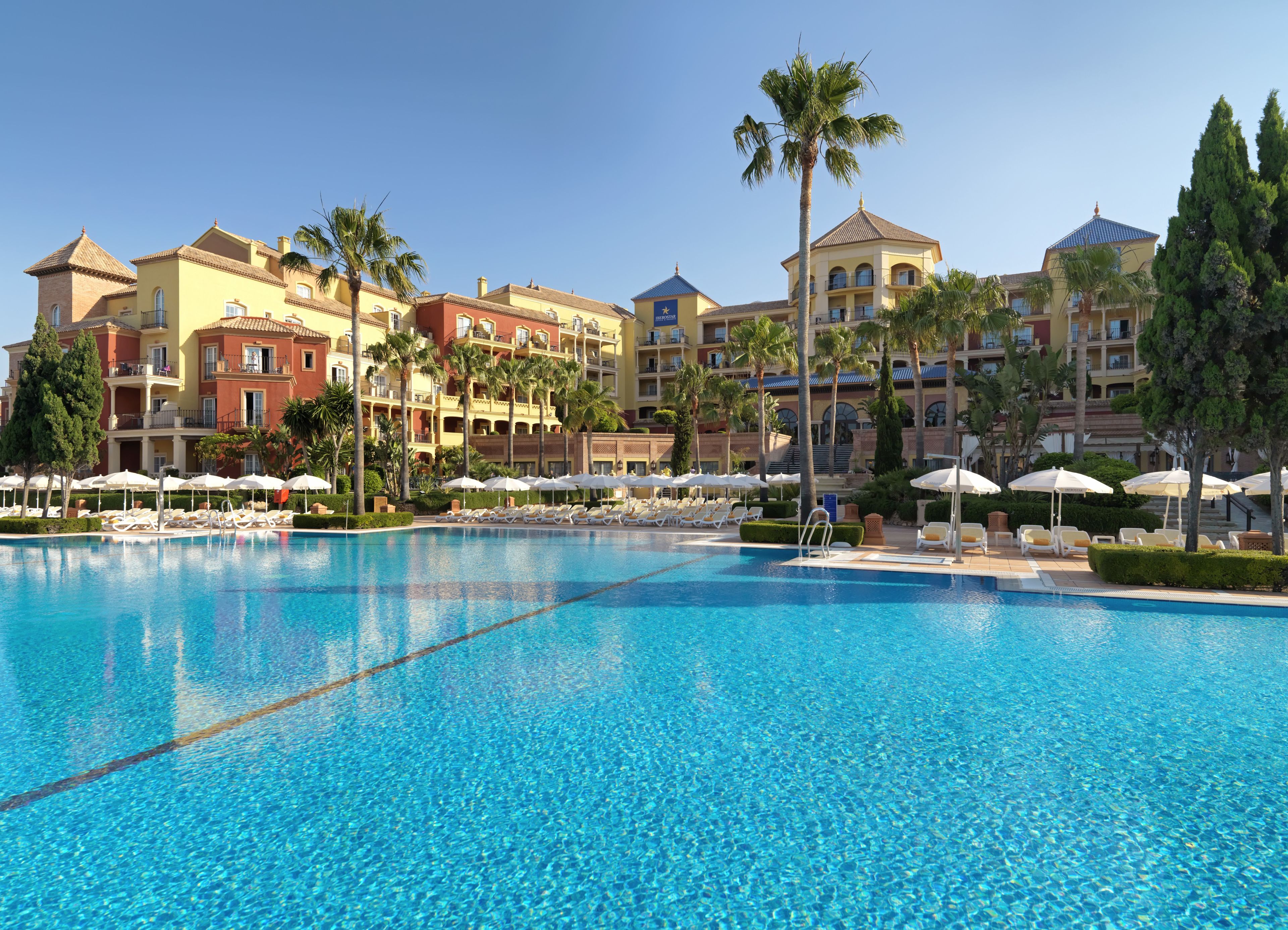 Una gran piscina con sillas de descanso y sombrillas, rodeada de edificios de hotel de varios pisos con balcones y palmeras.