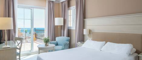 A hotel room with a large bed, a desk with a lamp, a chair, a small table with a plant, and a view of the sea through a balcony.