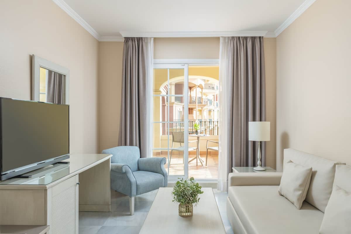 Quarto de hotel moderno com televisão de tela plana, sofá, poltrona, mesa de centro com uma planta e varanda com área de jantar.