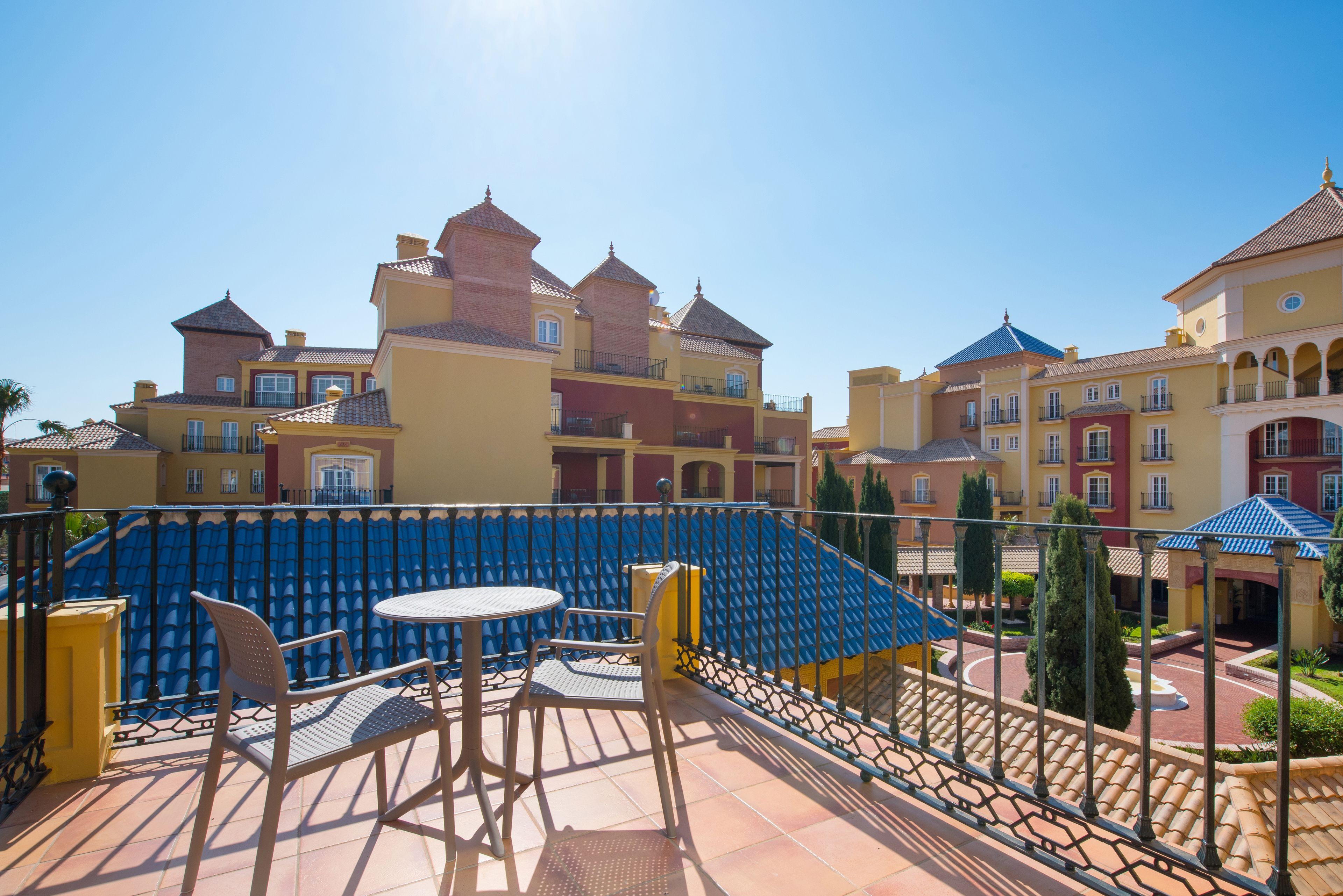 A balcony with a table and chairs, a blue railing, and a view of a multi-story building with balconies.