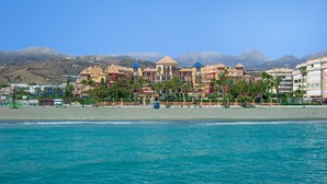 Ein Hotel direkt am Strand, mit mehreren Gebäuden, Palmen und Bergen im Hintergrund.