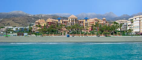 Un hotel frente a la playa con varias construcciones, palmeras y montañas al fondo.