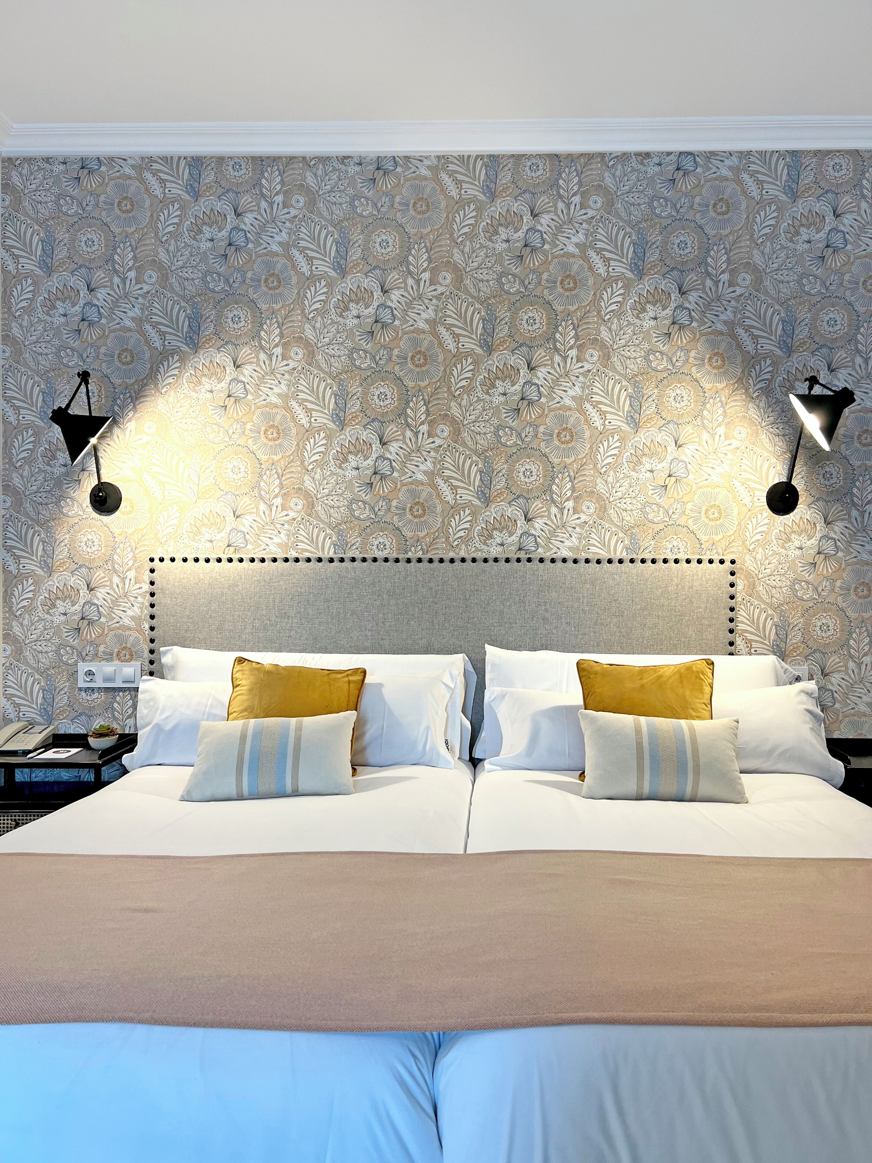 A neatly made bed with white and gold pillows, a beige throw blanket, and a headboard with a dotted pattern. Two black wall sconces provide lighting.