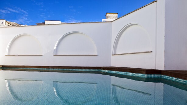 Una piscina con agua cristalina azul, rodeada de muros blancos con ventanas de arco.