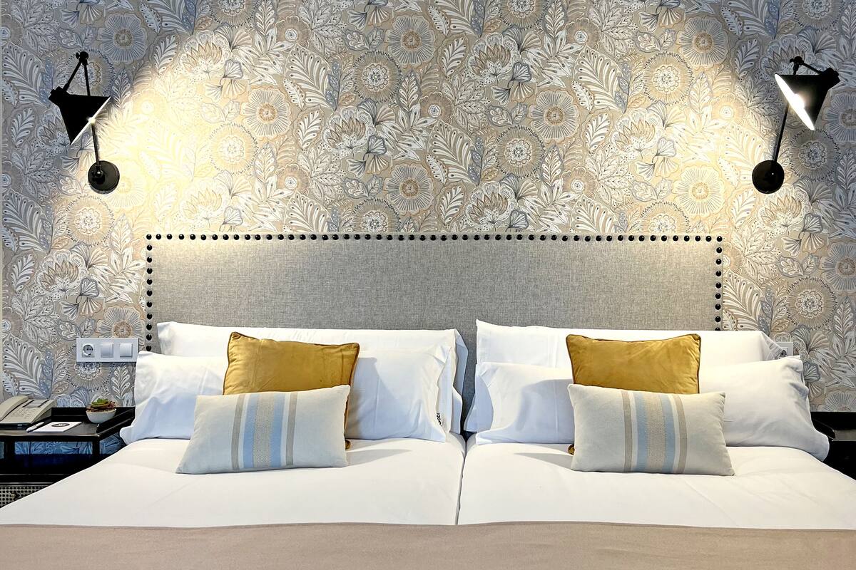 Un lit bien fait, agrémenté d’oreillers blancs et dorés, d’un plaid beige et d’une tête de lit à motifs à pois. Deux appliques murales noires assurent l’éclairage.