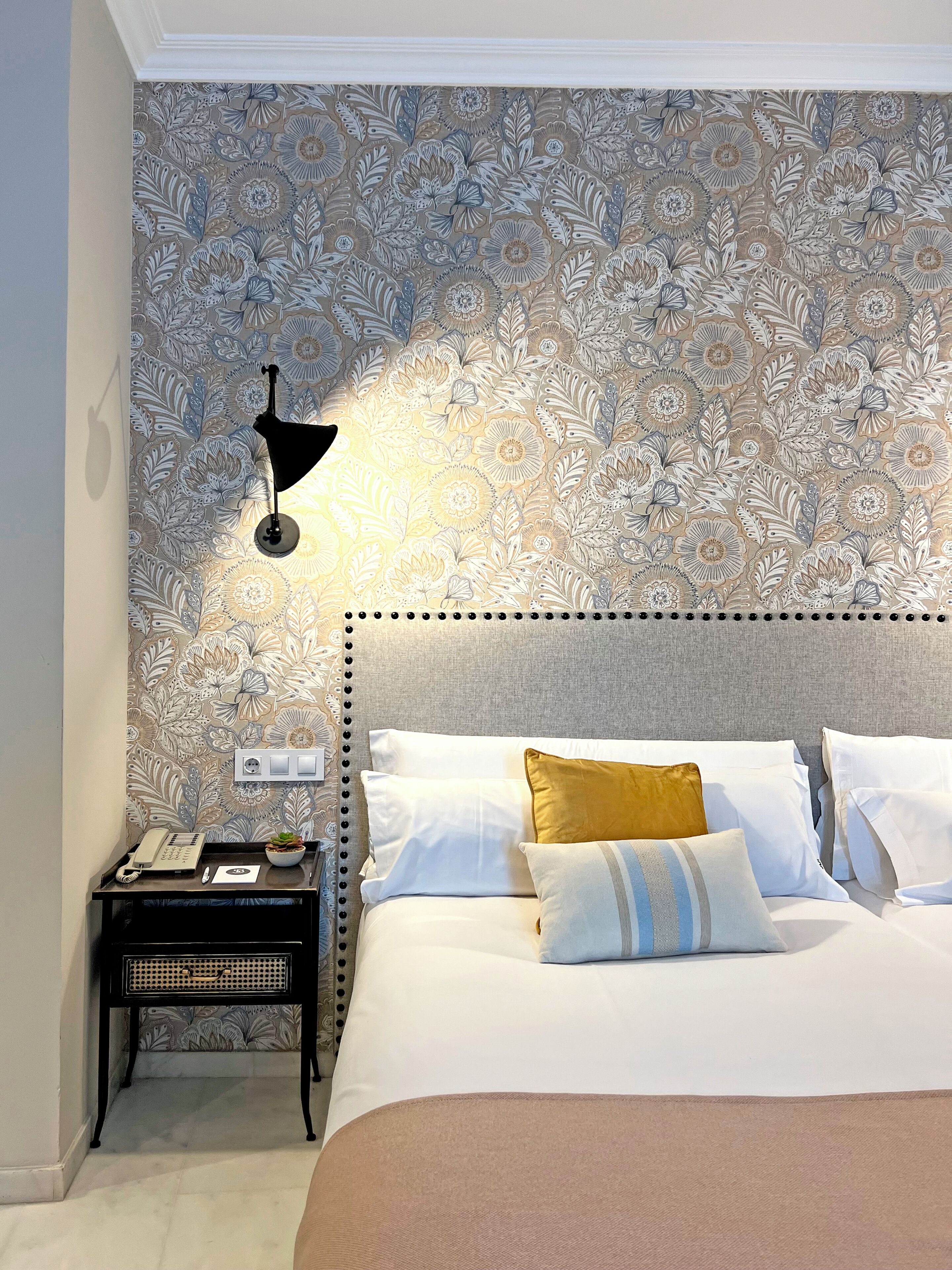 A bedroom with a patterned wallpaper, a bedside lamp, a nightstand with a phone, and a bed with pillows.