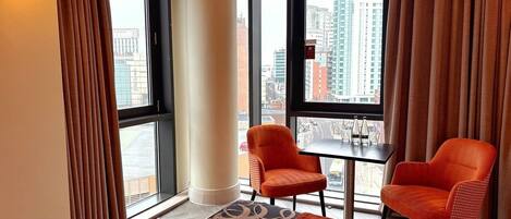 A hotel room with a large bed, two orange armchairs, a small table, and a view of the city through large windows.