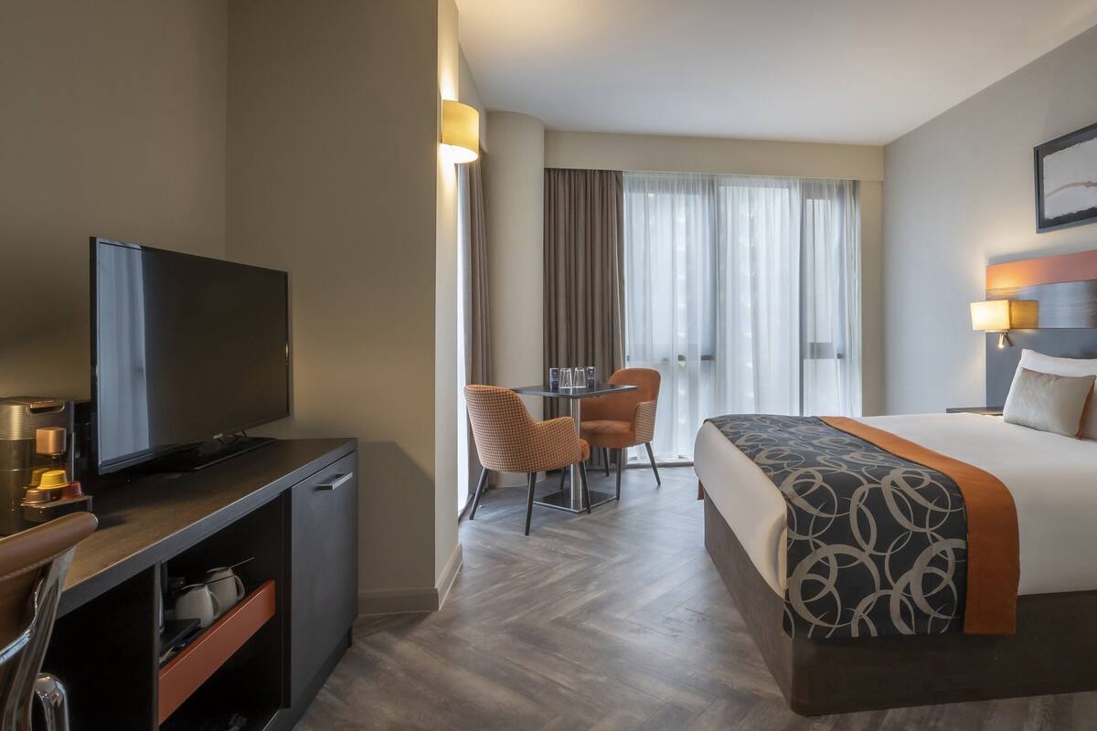 A hotel room with a bed, a TV, a desk with chairs, and a window with curtains.