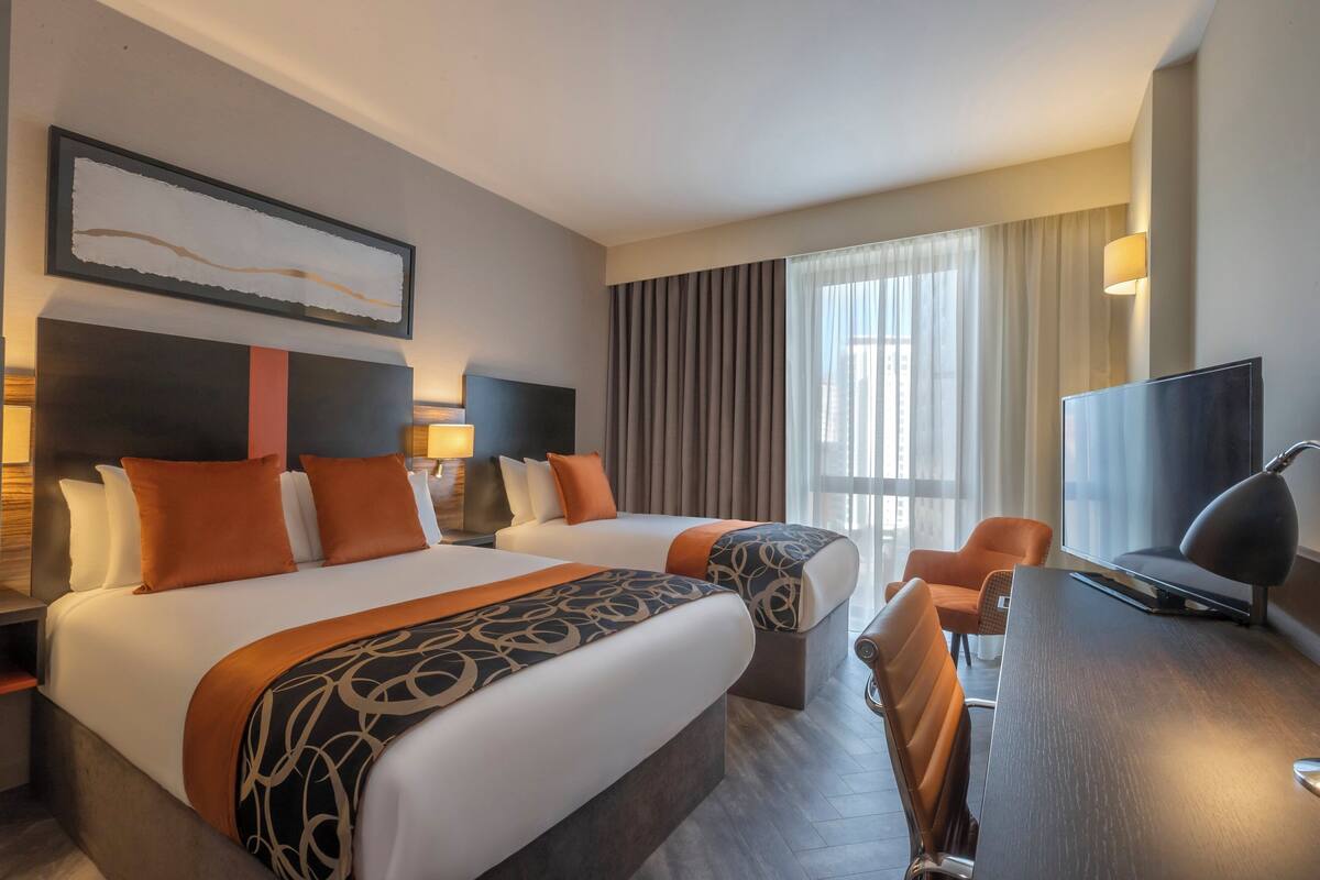 A hotel room with two beds, a desk, a TV, and a large window with curtains.