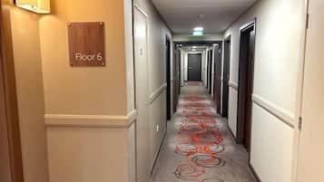 Ein Hotelgang mit Teppich, der rote, kreisförmige Muster aufweist, Türen an beiden Seiten und einem Schild mit der Aufschrift '6. Stockwerk'.