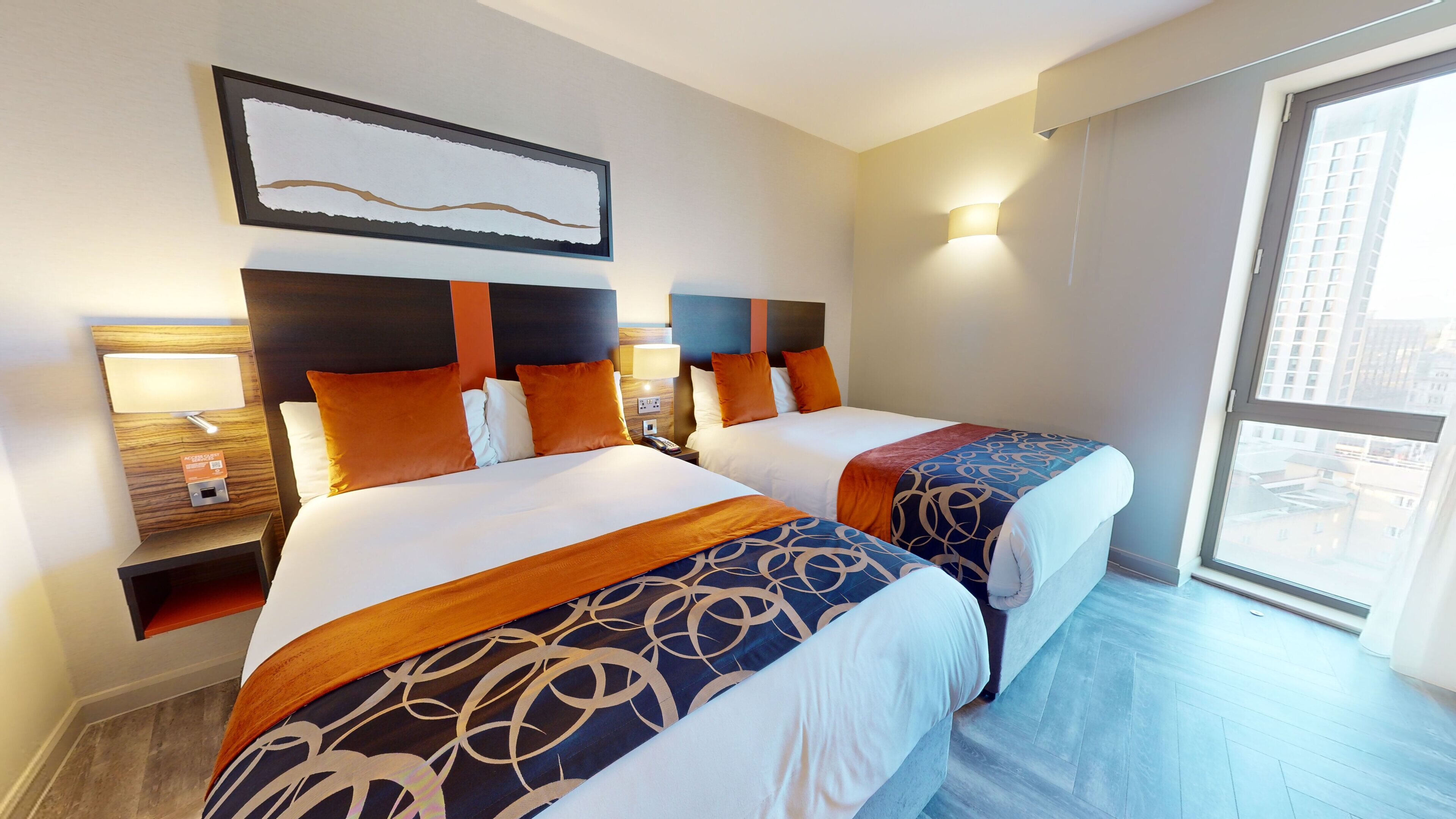 Hotellihuone, jossa on kaksi sänkyä, yöpöytä, lamppu ja suuri ikkuna, josta on näkymä kaupunkiin.